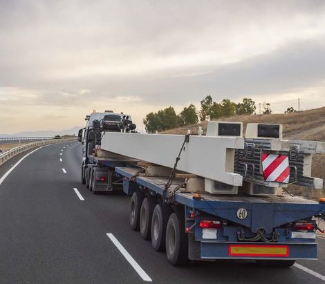 Camión que transporta una larga viga de hormigón por una autopista, con una señal de peligro roja y blanca en la parte trasera.