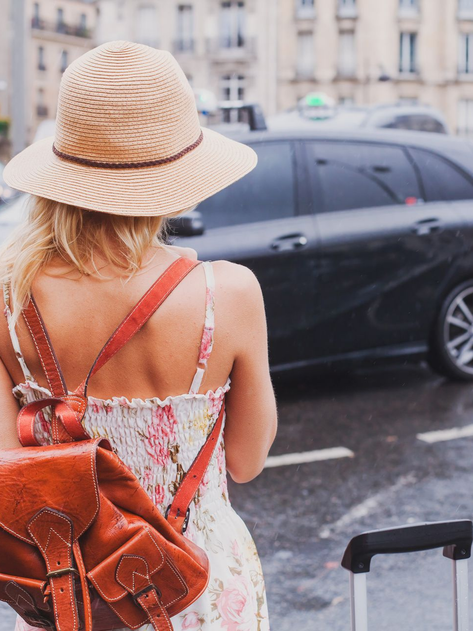 Une femme avec un chapeau et un sac à dos attendant près d'un taxi, tenant une valise dans une rue de la ville.