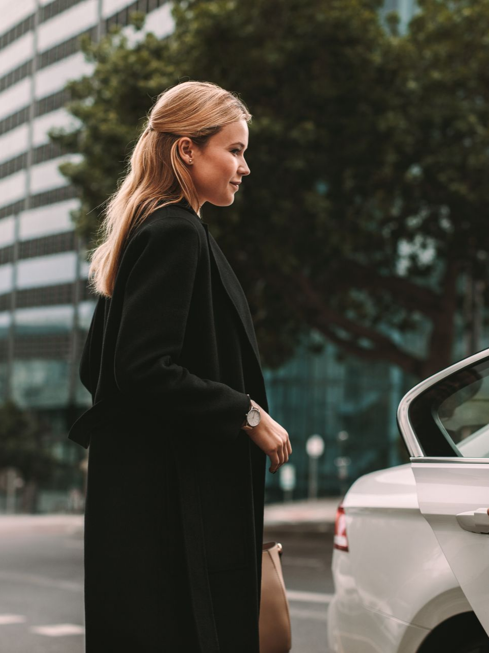 Femme en manteau noir près d'une voiture, arrière-plan de la ville.