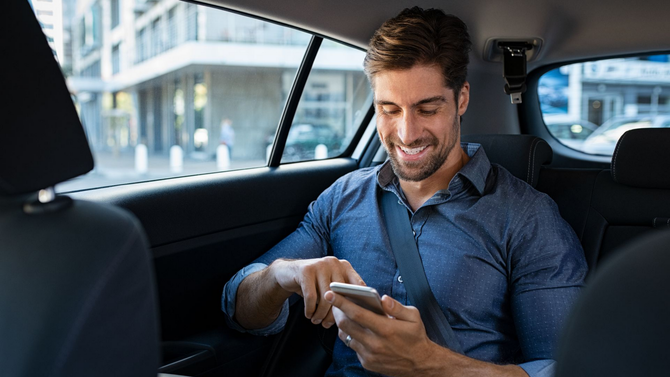 Un homme en voiture sourit à son téléphone. Il porte une chemise bleue et porte sa ceinture de sécurité.
