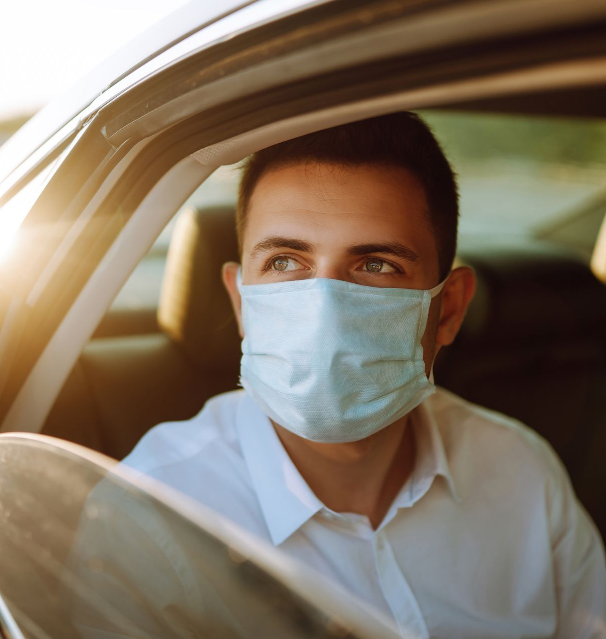 Un homme portant un masque facial dans une voiture, regardant par la fenêtre ; la lumière du soleil brille à l'intérieur.