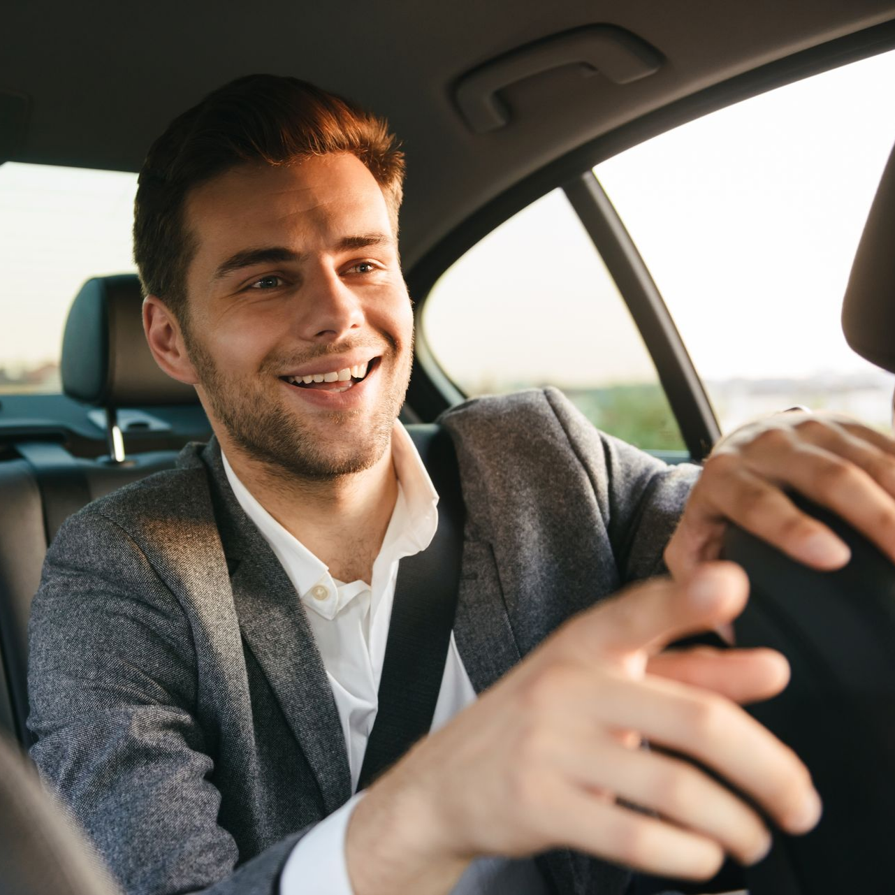 Un homme souriant sur le siège arrière d'une voiture, portant une veste de costume et une ceinture de sécurité.