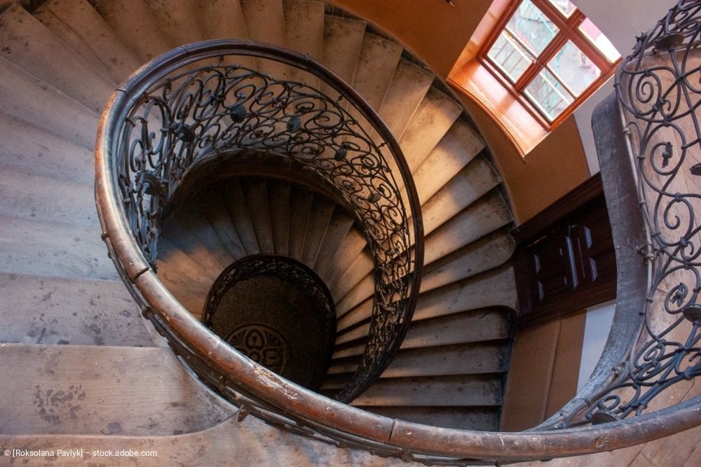 Eine Ansicht von oben nach unten auf eine gewundene Steintreppe mit einem kunstvollen, schwarzen schmiedeeisernen Geländer.