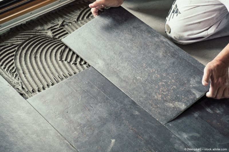 Ein Arbeiter legt eine dunkelgraue, rechteckige Bodenfliese auf einen mit Klebstoff bedeckten Betonboden.