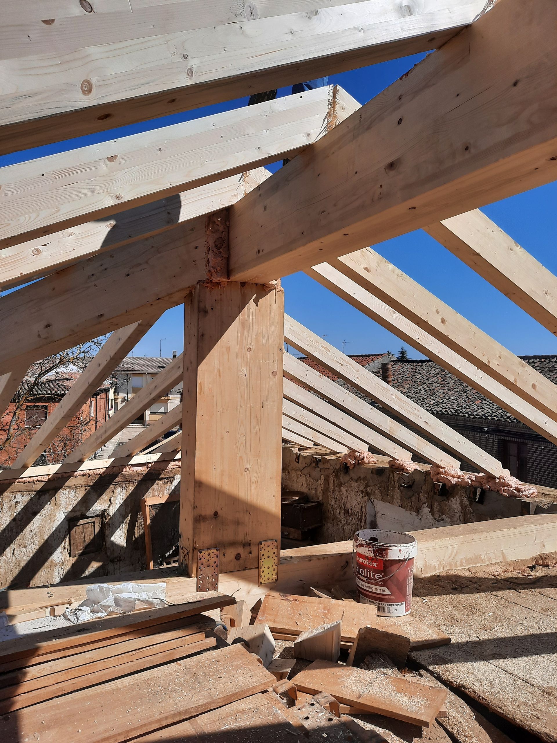 Estructura de techo de madera en construcción, con vigas vistas y un poste de soporte central. Cielo azul brillante.