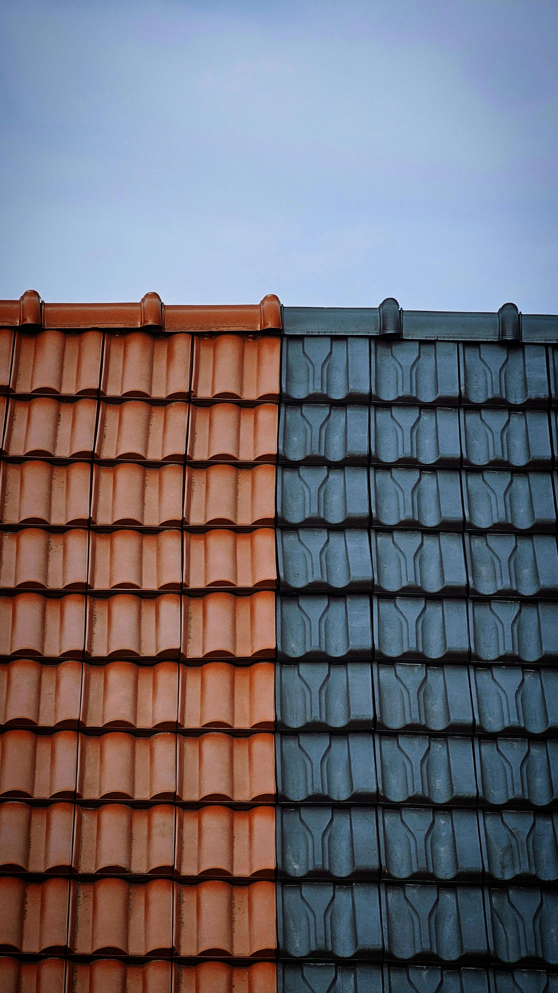 Techo de dos tonos con tejas naranjas y azules contra un cielo azul pálido.