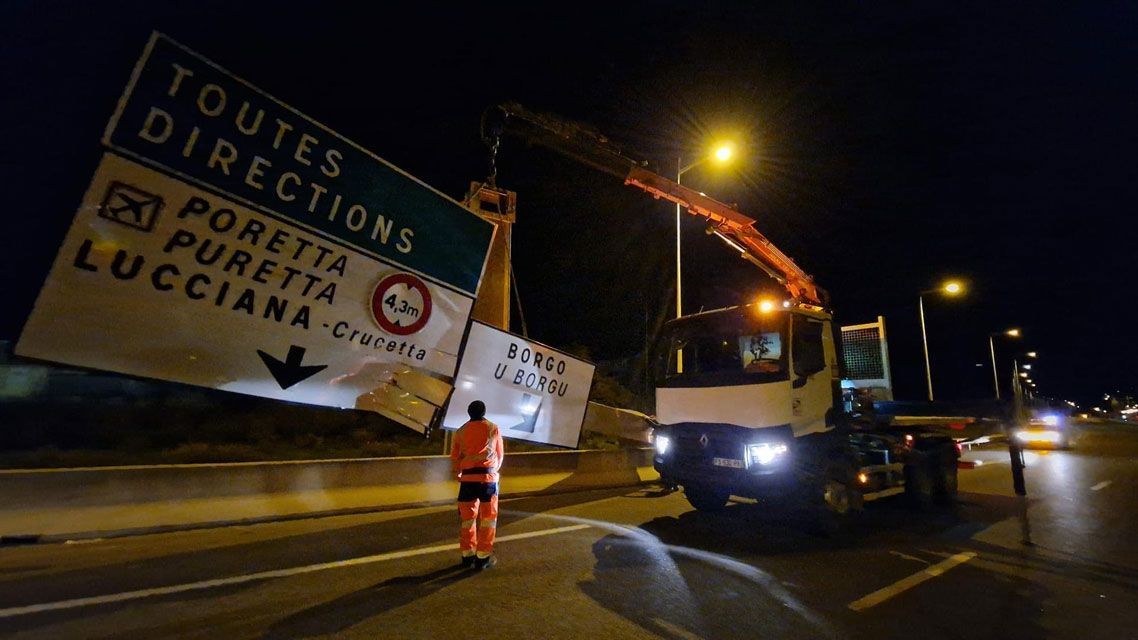 Un ouvrier en gilet orange guide une grue qui soulève un panneau de signalisation tombé, de nuit, sur une chaussée mouillée.