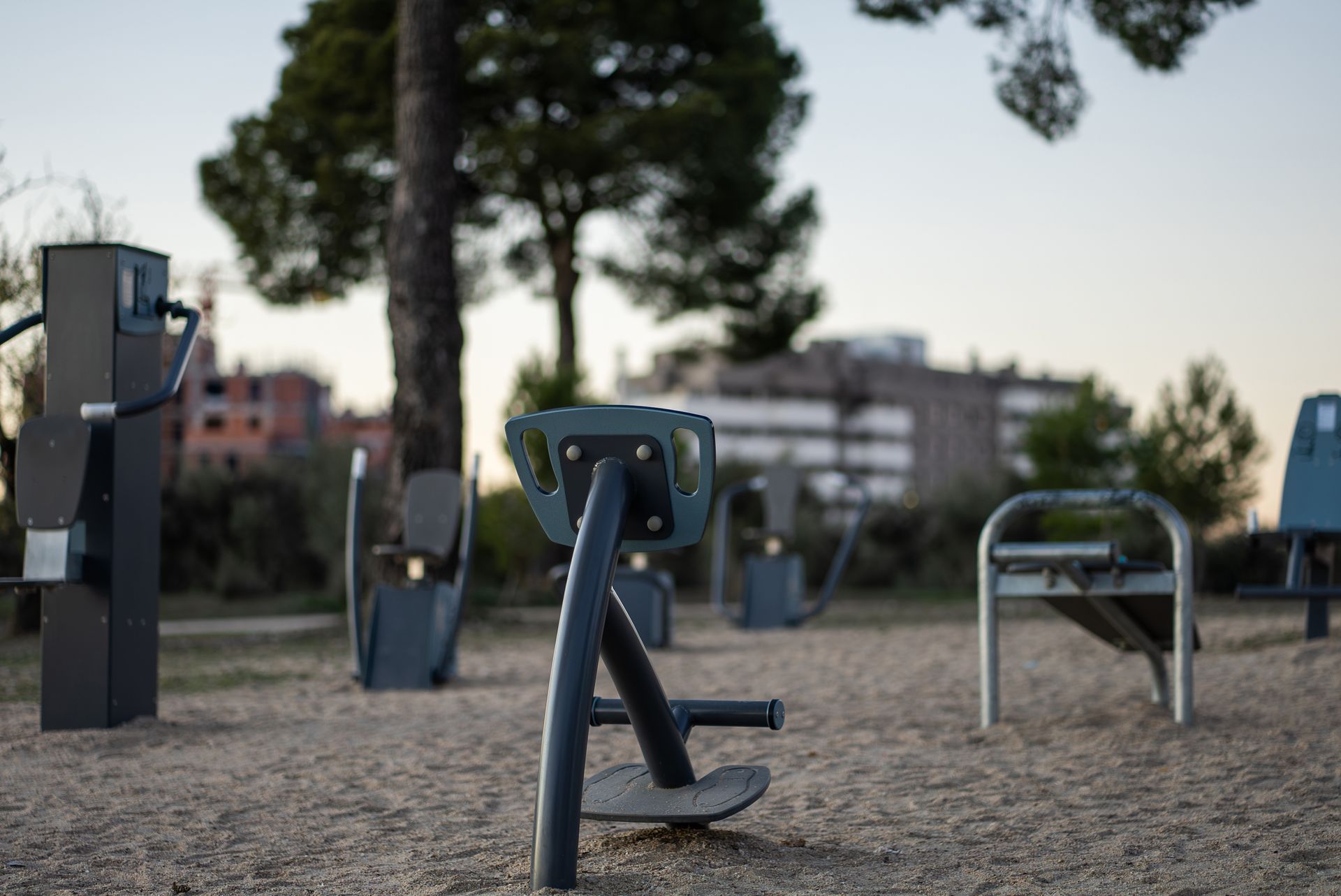 Équipements de fitness en plein air sur une surface gravillonnée dans un parc, avec des arbres et des bâtiments en arrière-plan.