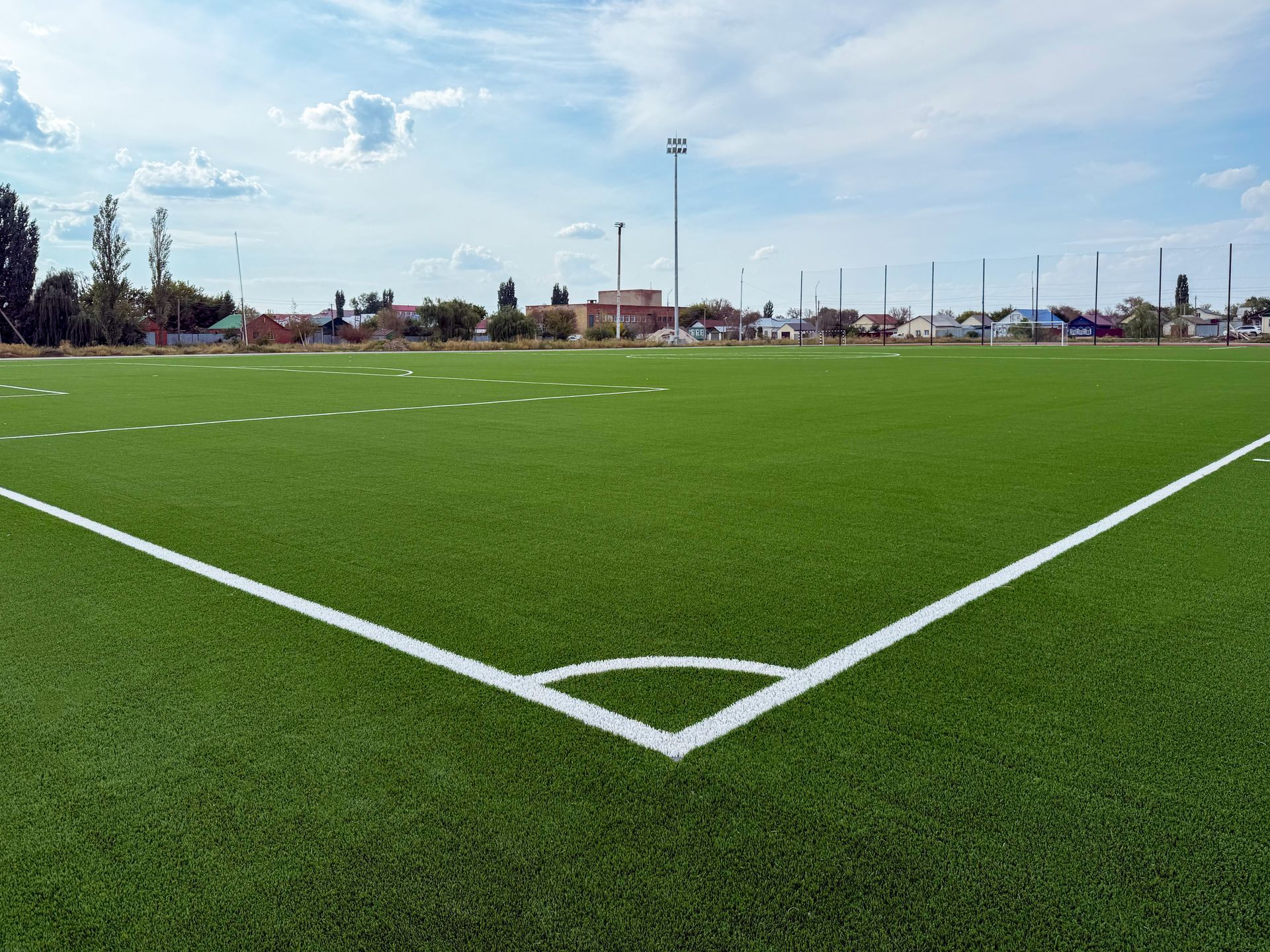 Un coin de terrain de football avec des lignes blanches délimitant un gazon synthétique vert vif, sous un ciel bleu nuageux.