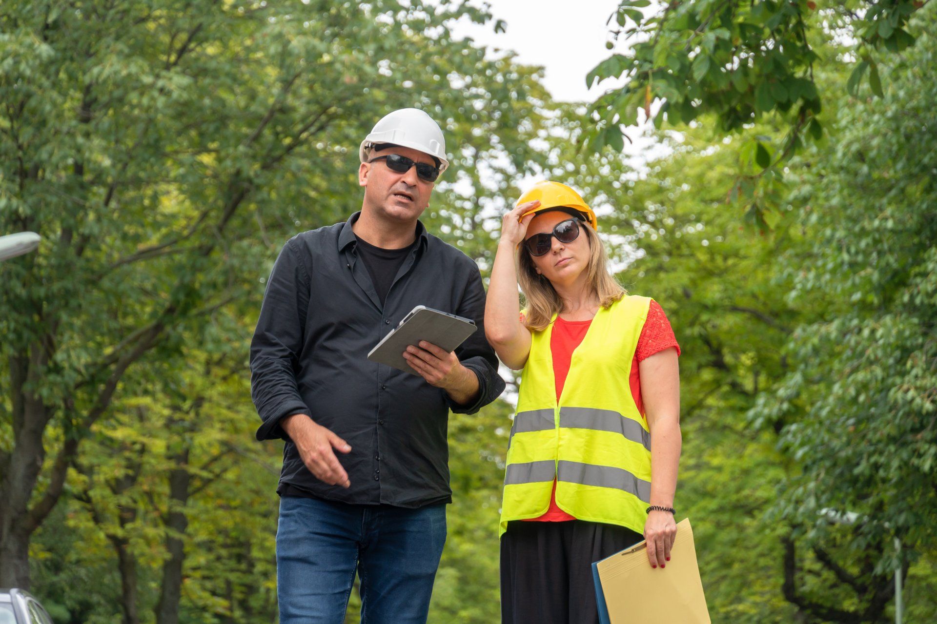 Deux personnes portant des casques de chantier et des lunettes de soleil se tiennent à l'extérieur, l'une tenant une tablette et l'autre un dossier.