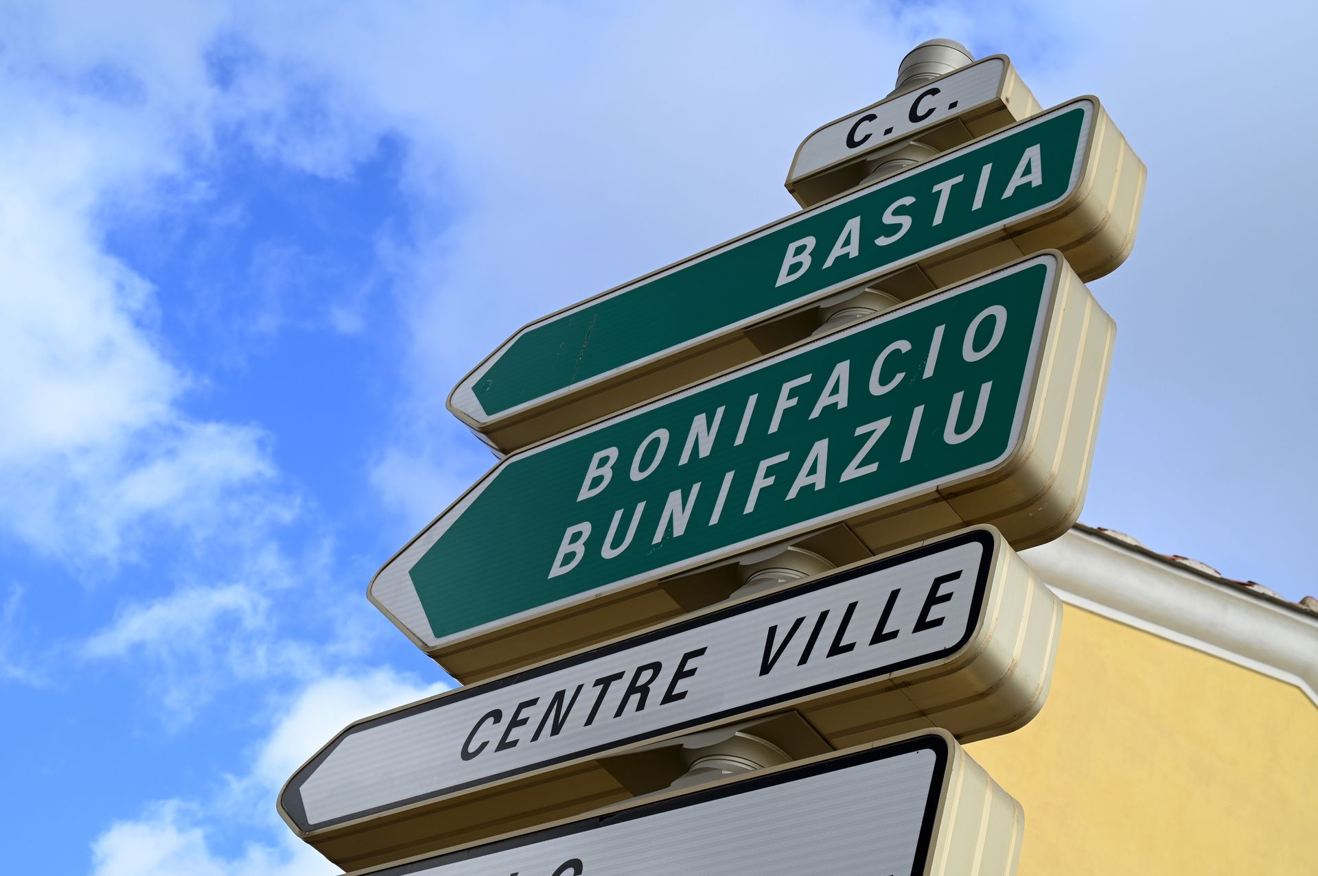 Panneaux routiers sur un poteau se détachant sur un ciel bleu, indiquant Bastia, Bonifacio/Bunifaziu et le centre-ville.