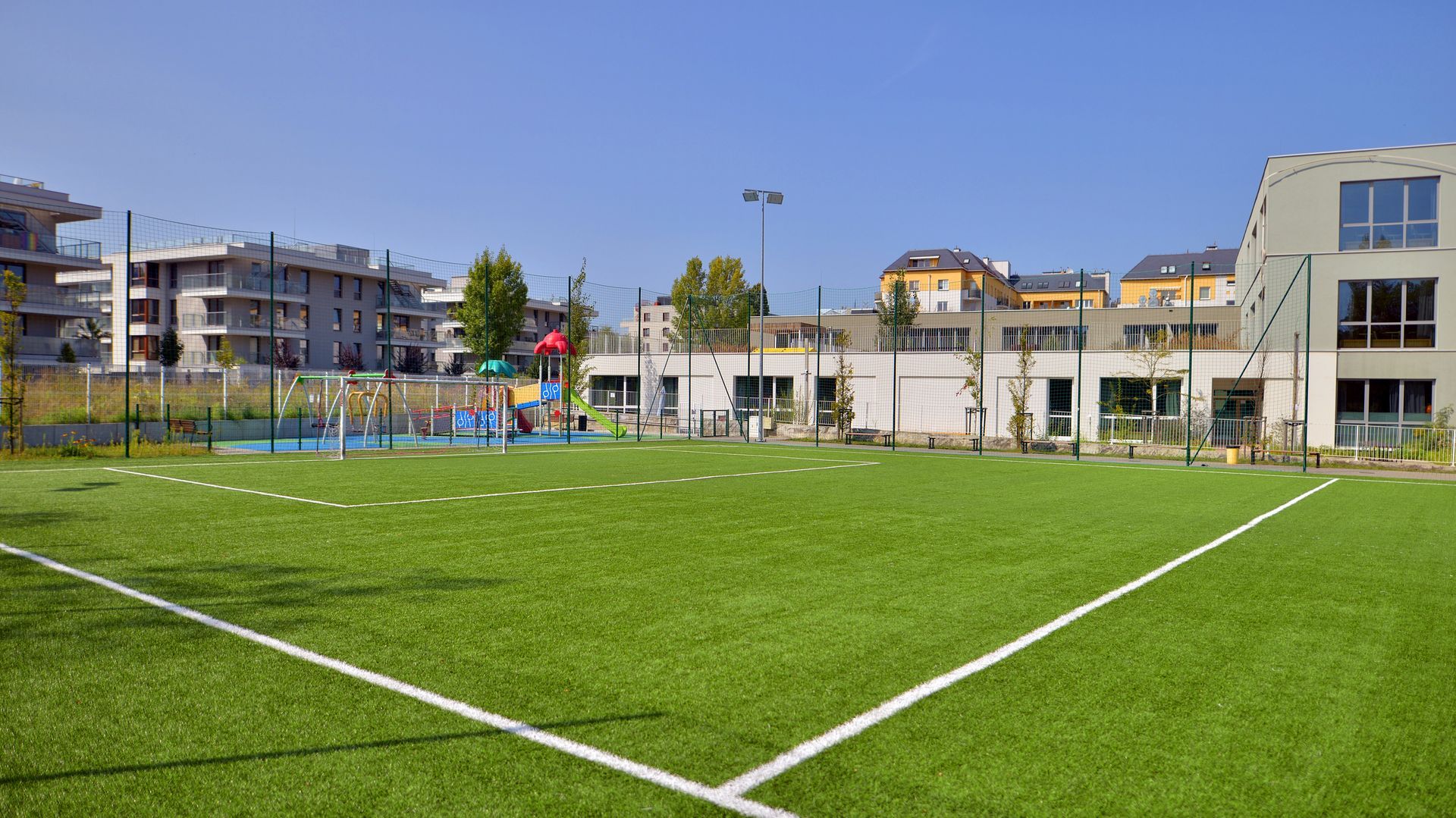 Un terrain de football avec une pelouse artificielle d'un vert éclatant, des lignes blanches délimitant le terrain et des bâtiments résidentiels tout autour.