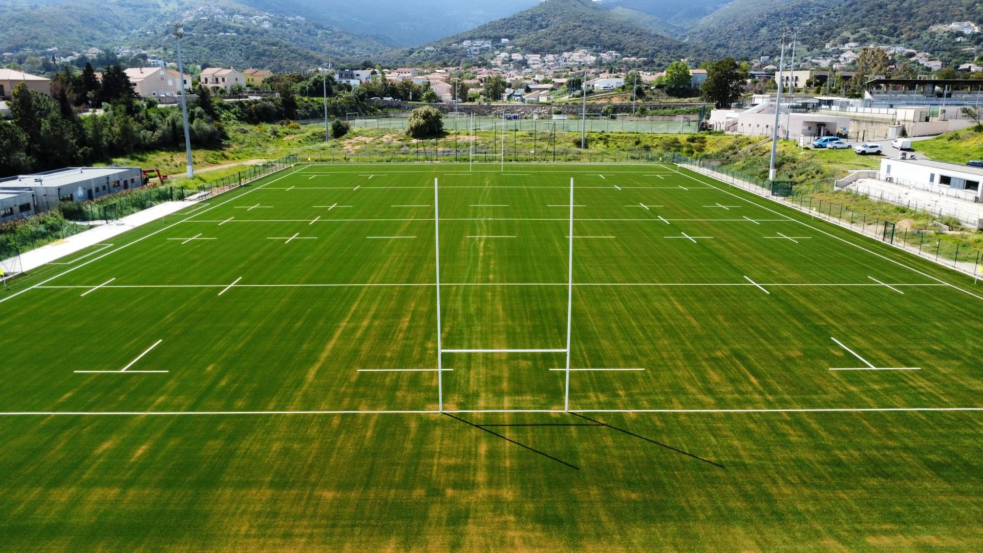 Terrain de rugby vide avec des poteaux de but sur une pelouse verte, montagnes et bâtiments en arrière-plan