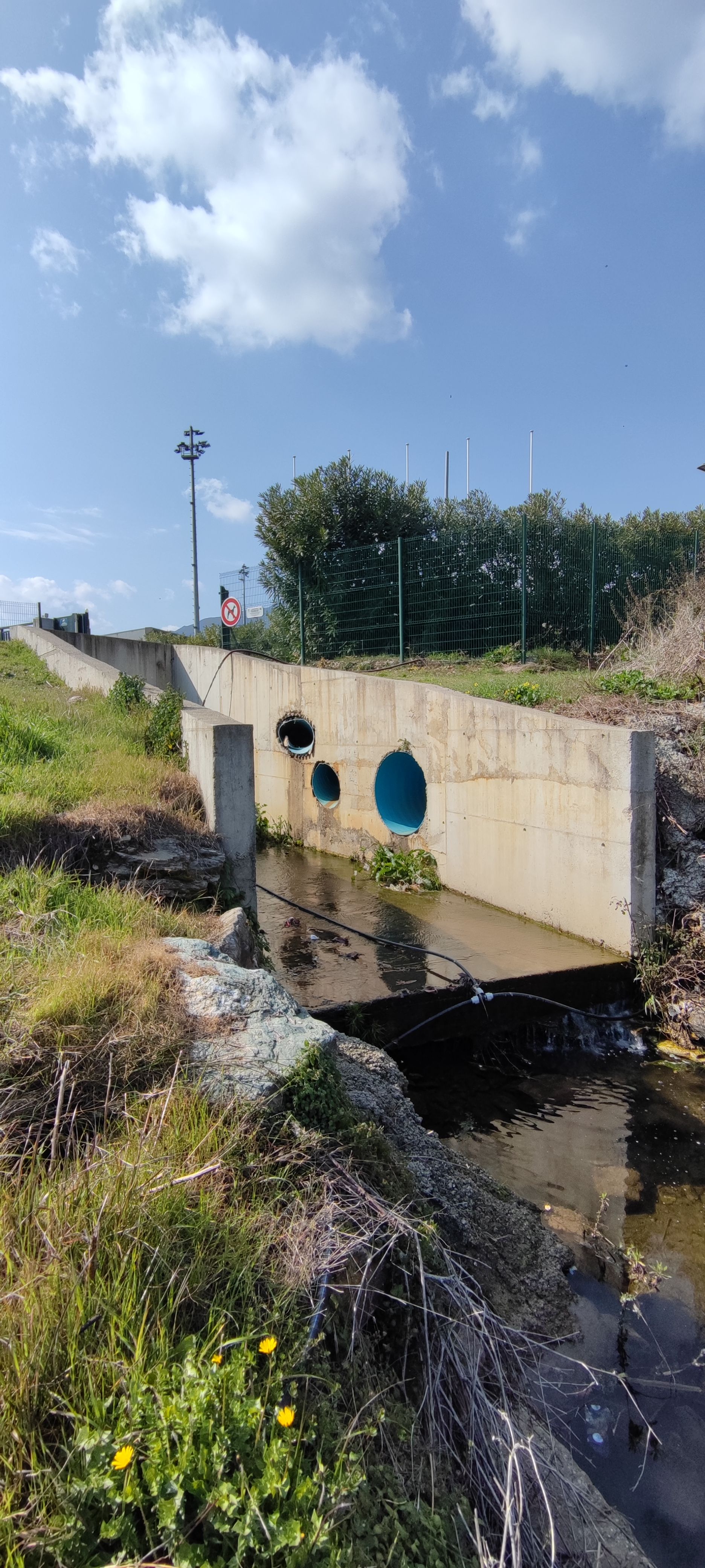 Pont en béton enjambant un ruisseau étroit bordé de végétation verdoyante sous un ciel bleu nuageux