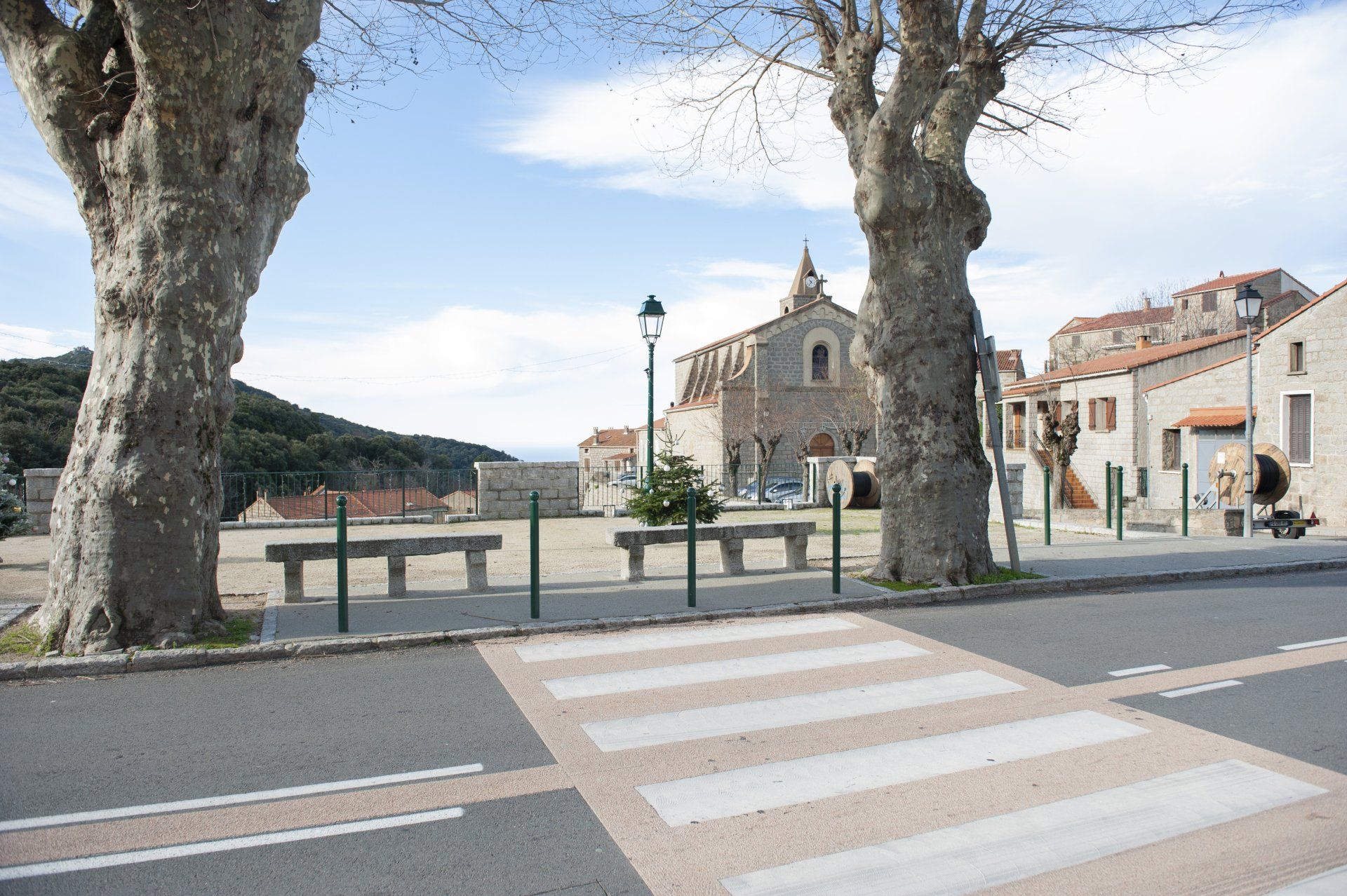 Un passage piéton mène à une place de village ensoleillée, bordée de bâtiments en pierre, de bancs et de grands arbres sous un ciel d'un bleu éclatant.