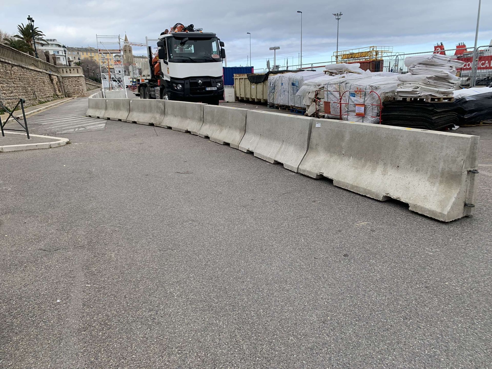 Une rangée de barrières Jersey en béton est posée sur une surface asphaltée près d'un camion blanc et d'une pile de matériaux palettisés.