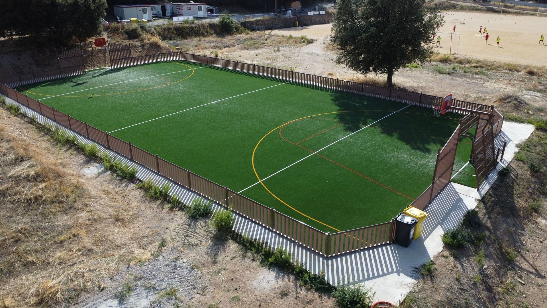 Un terrain de football extérieur clôturé, recouvert de gazon synthétique, avec des marquages blancs et jaunes et des paniers de basket.