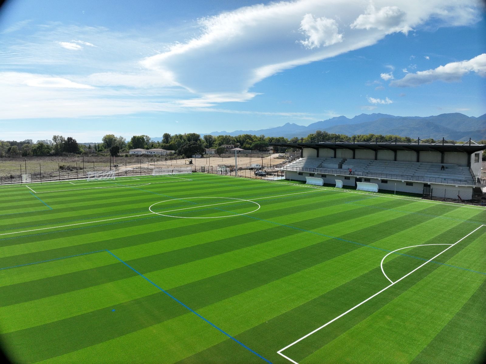 Vue aérienne d'un terrain de football d'un vert éclatant sous un ciel bleu ensoleillé, avec des montagnes en arrière-plan.