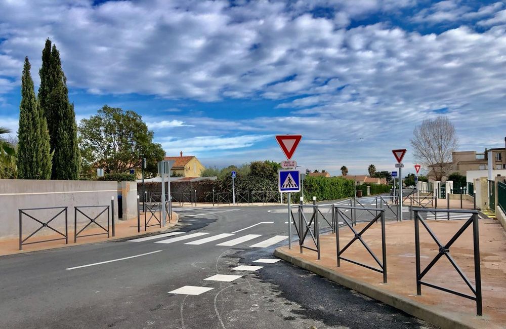Une rue tranquille de banlieue avec un passage piéton, des panneaux « cédez le passage », des barrières métalliques et des arbres sous un ciel bleu nuageux.