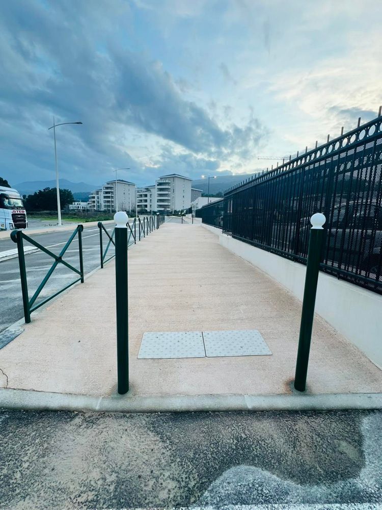 Une allée piétonne pavée, entre une clôture métallique et une rambarde verte, menant vers des immeubles résidentiels au crépuscule.