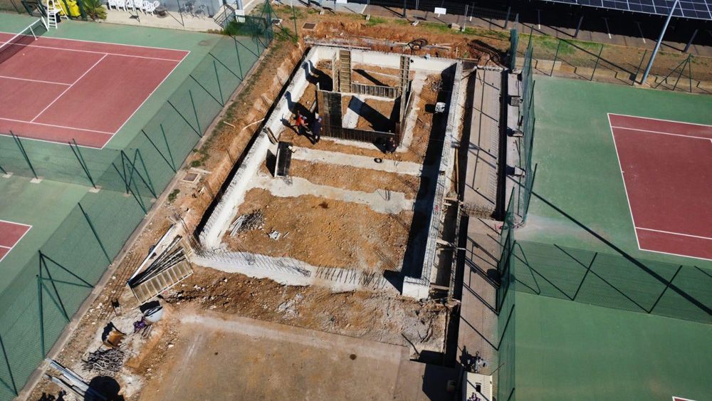 Vue aérienne d'un chantier de construction à côté de courts de tennis, avec des fondations en béton et de la terre excavée.