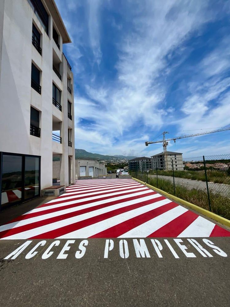 Une voie d'accès pour les pompiers, délimitée par des bandes rouges et blanches sur le trottoir, longe un immeuble d'appartements blanc.