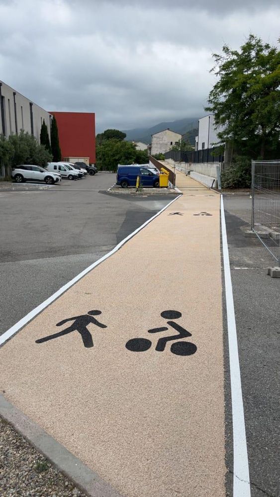 Un chemin partagé beige et pavé, balisé par des icônes noires, pour les piétons et les cyclistes, traverse un parking extérieur.