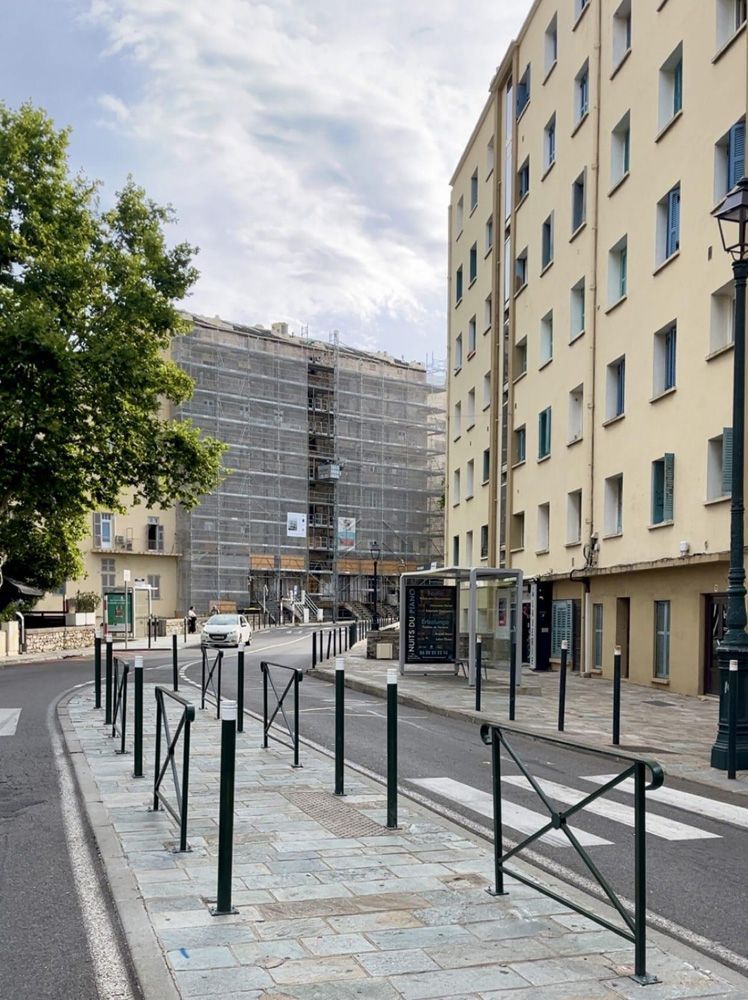 Une scène de rue en ville, avec un grand immeuble jaune clair à droite et un bâtiment en travaux à l'arrière-plan.