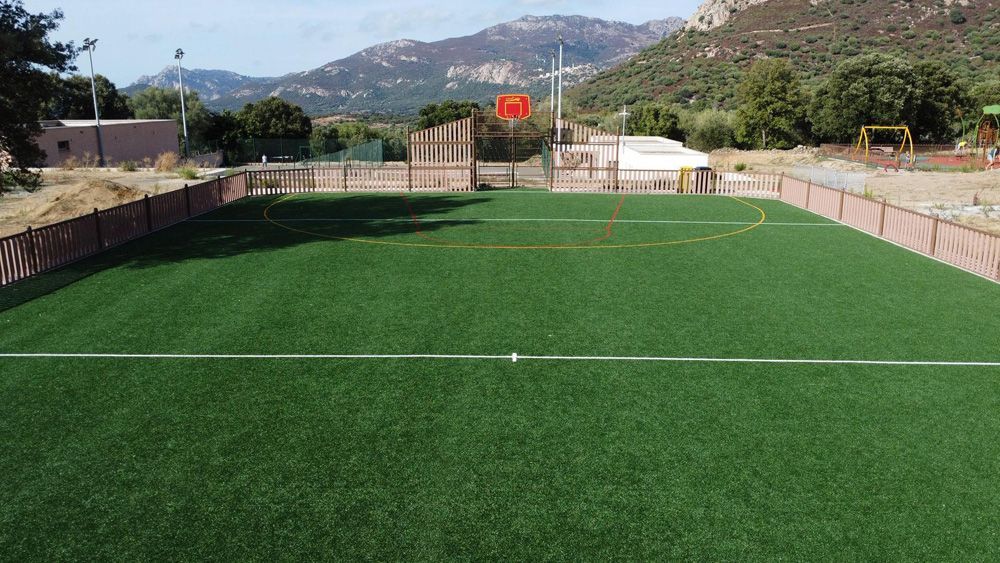 Petit terrain de football extérieur avec gazon vert, montagnes en arrière-plan et drapeau rouge au centre.
