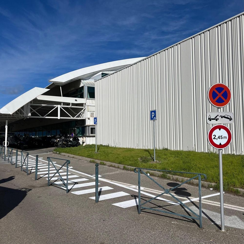 Un bâtiment d'aéroport avec un parking, une signalétique et un passage piéton par une journée ensoleillée.