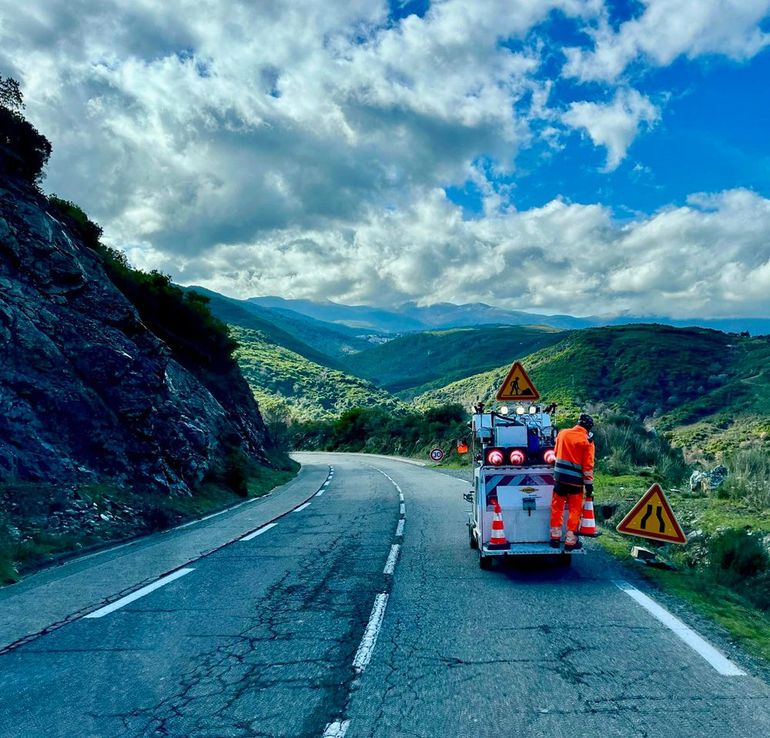 Une équipe de travaux publics travaille sur une route de montagne sinueuse, avec des panneaux de signalisation et un camion utilitaire sous un ciel nuageux.