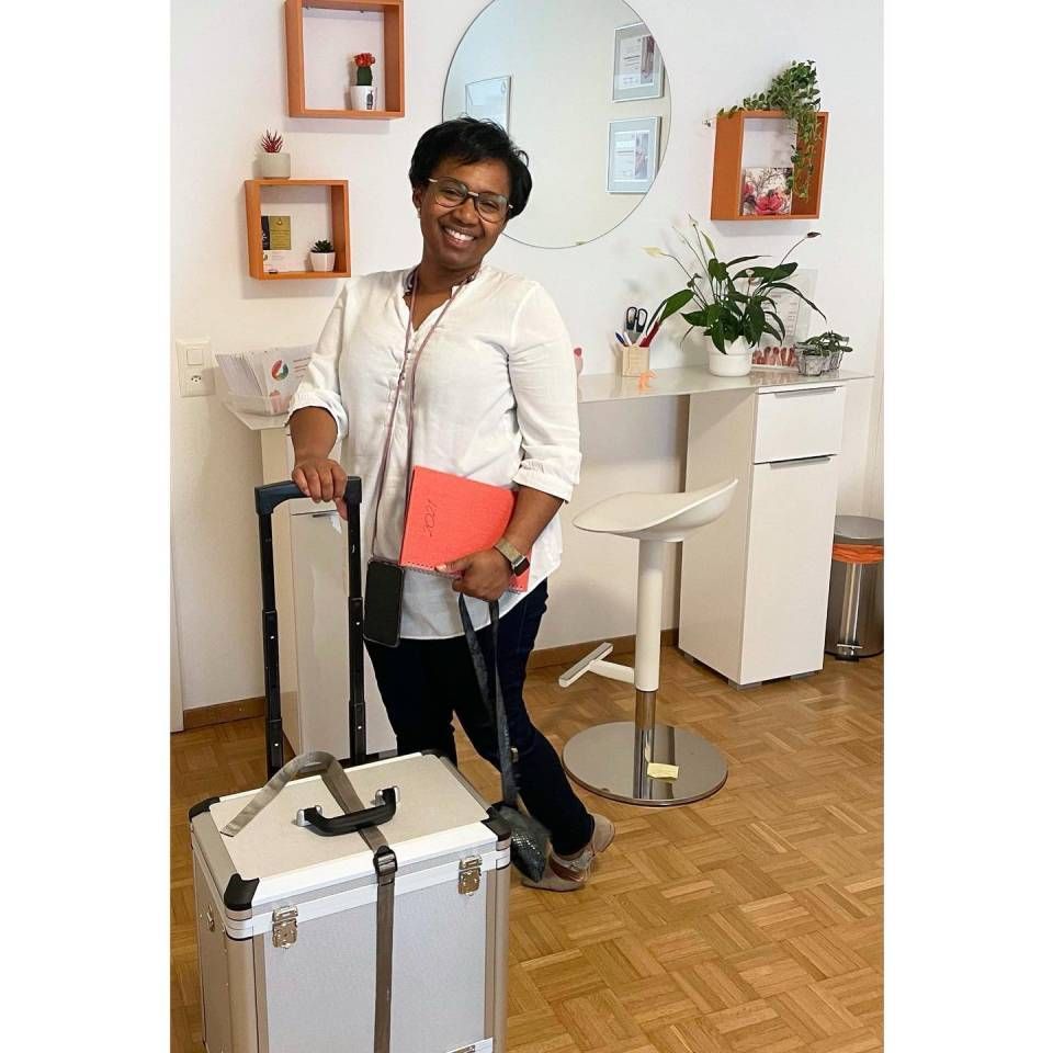 Une femme se tient debout à côté d'une valise à roulettes dans un bureau à domicile lumineux, tenant un dossier et souriant.