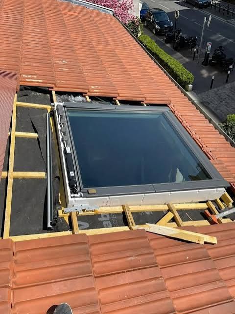 Installation d'une lucarne sur un toit en tuiles de terre cuite, avec cadre apparent et matériaux de construction à proximité.