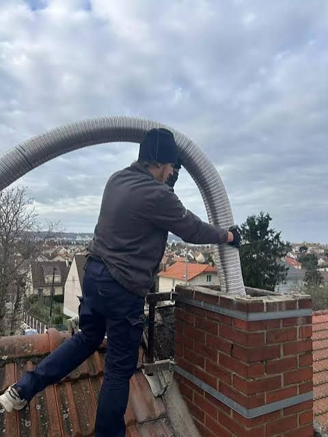 Une personne sur un toit utilise un grand tuyau flexible près d'une cheminée en briques, avec un ciel nuageux en arrière-plan.