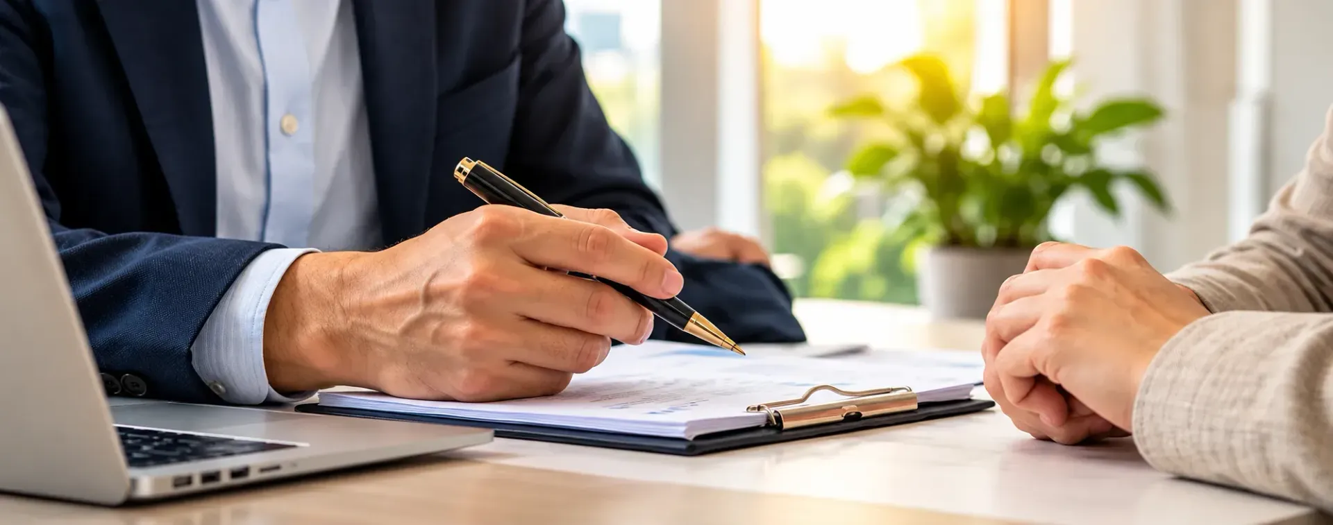 Person in suit signing documents across from another person at a desk in a bright office
