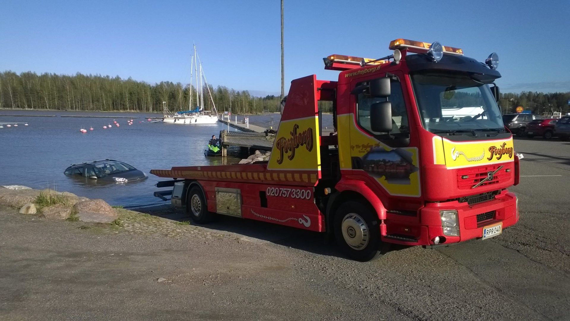 Punainen hinausauto pysäköitynä järvenrantatielle pienen veneen viereen vedessä
