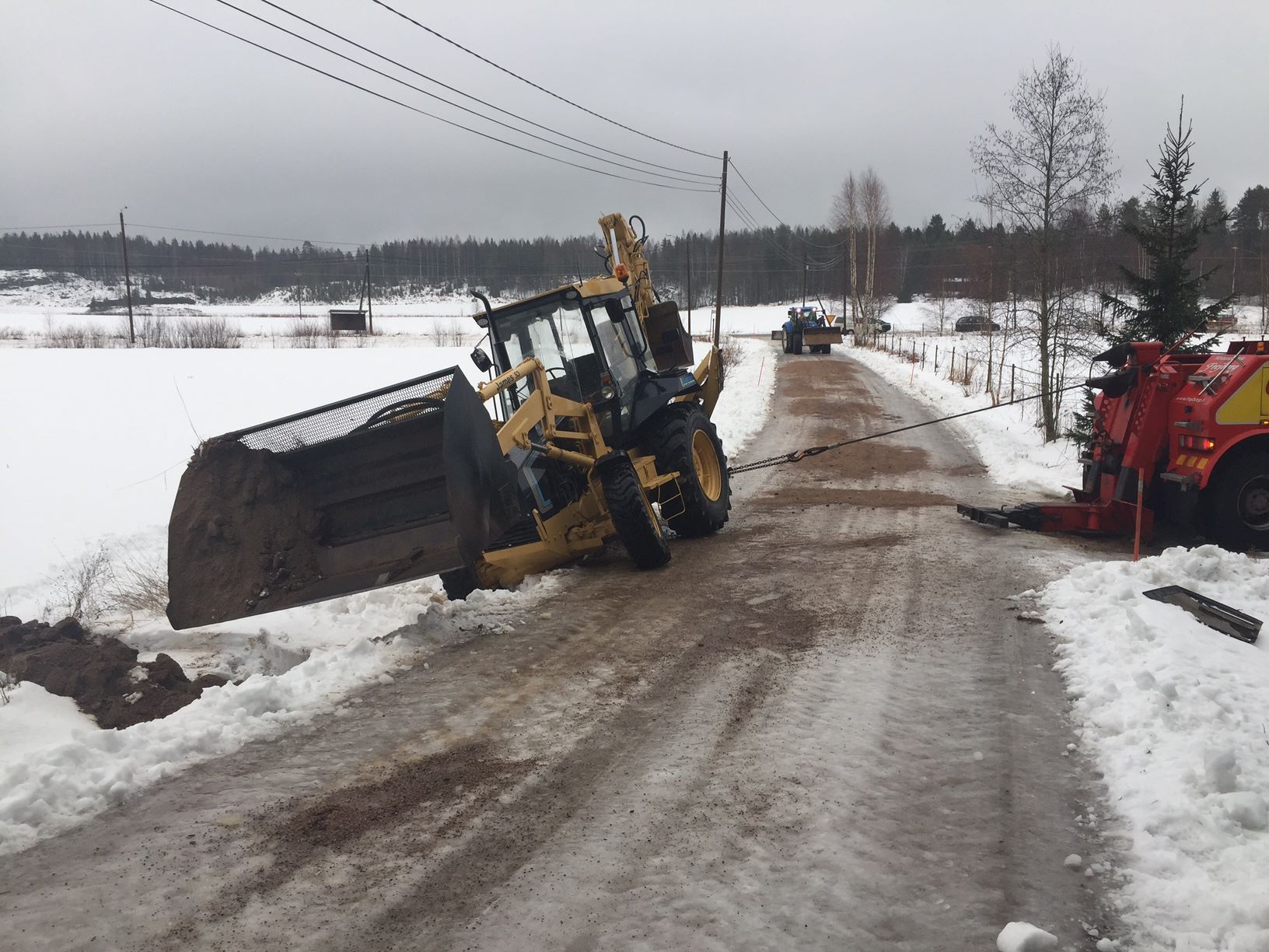 Keltainen kuormaaja kaadettuna lumisella tiellä kauha ylhäällä; punainen laite on tien laidassa.