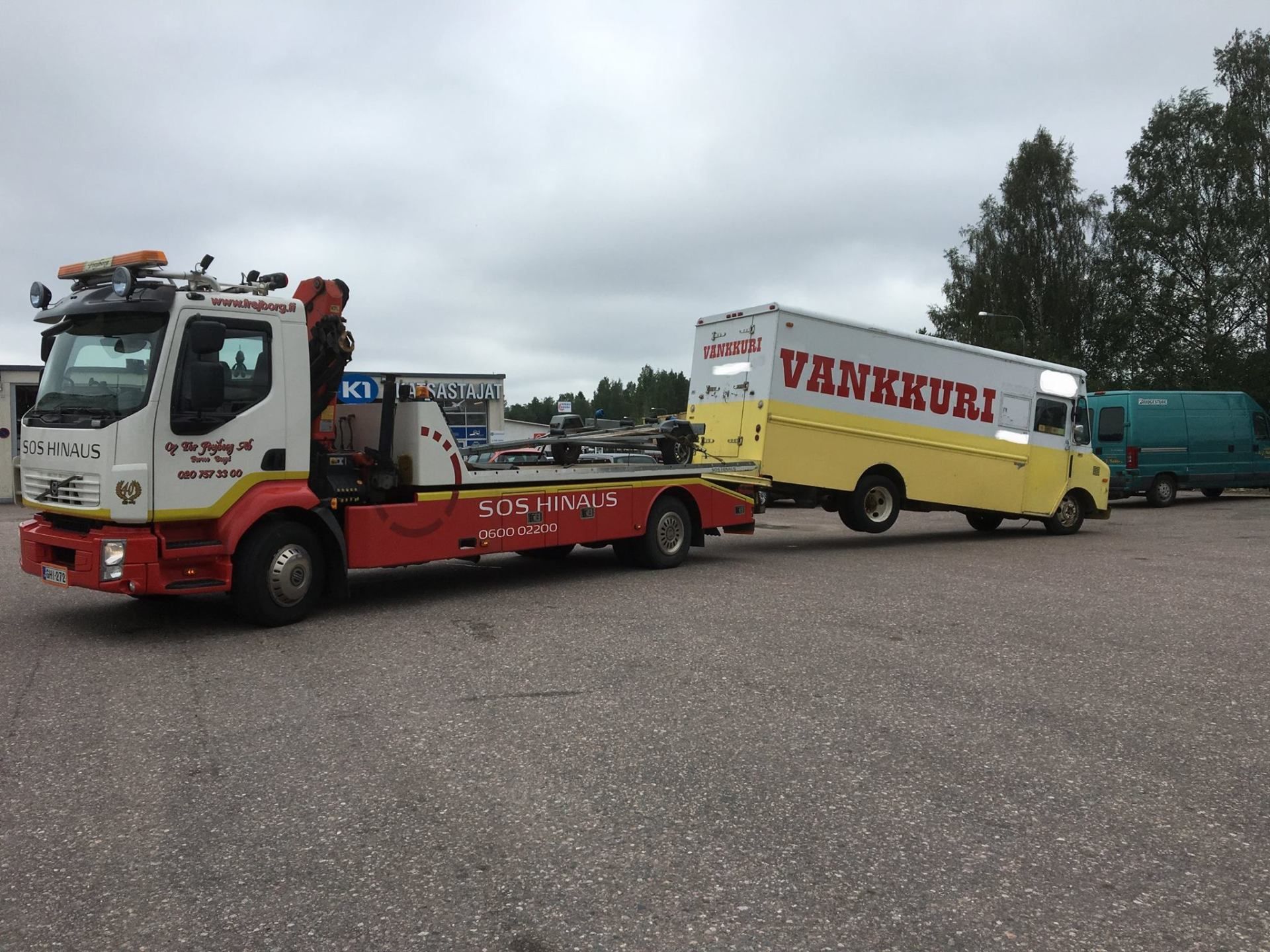 Punainen hinausauto vetää keltaista VÄNKKÄRI-perävaunua pysäköintialueella pilvisen taivaan alla