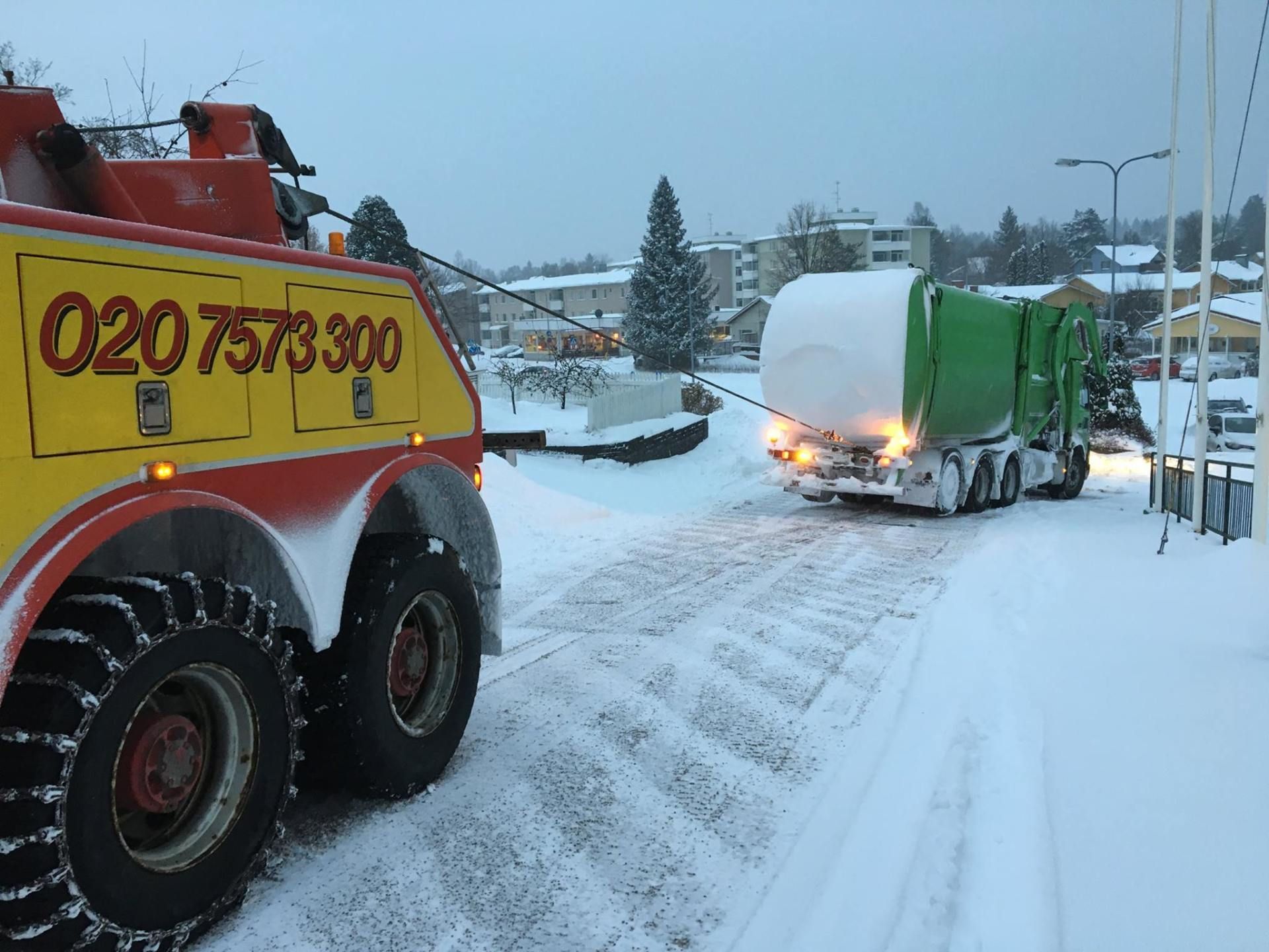 Hinausauto ja vihreä säiliöauto jumissa lumisella kadulla, renkaanjäljet lumessa