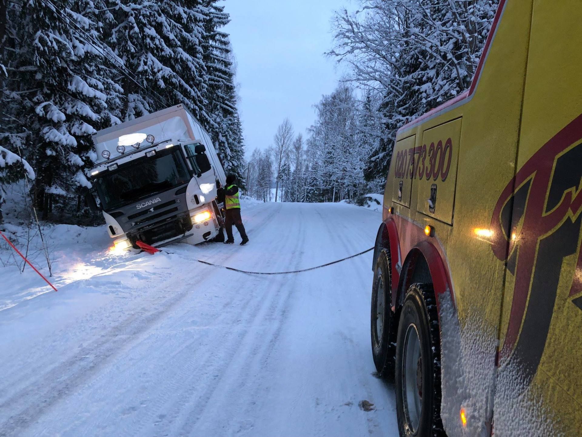 Hinausauto vetää lumen juuttunutta kuorma-autoa lumisella tiellä, etualalla keltainen hinausauto