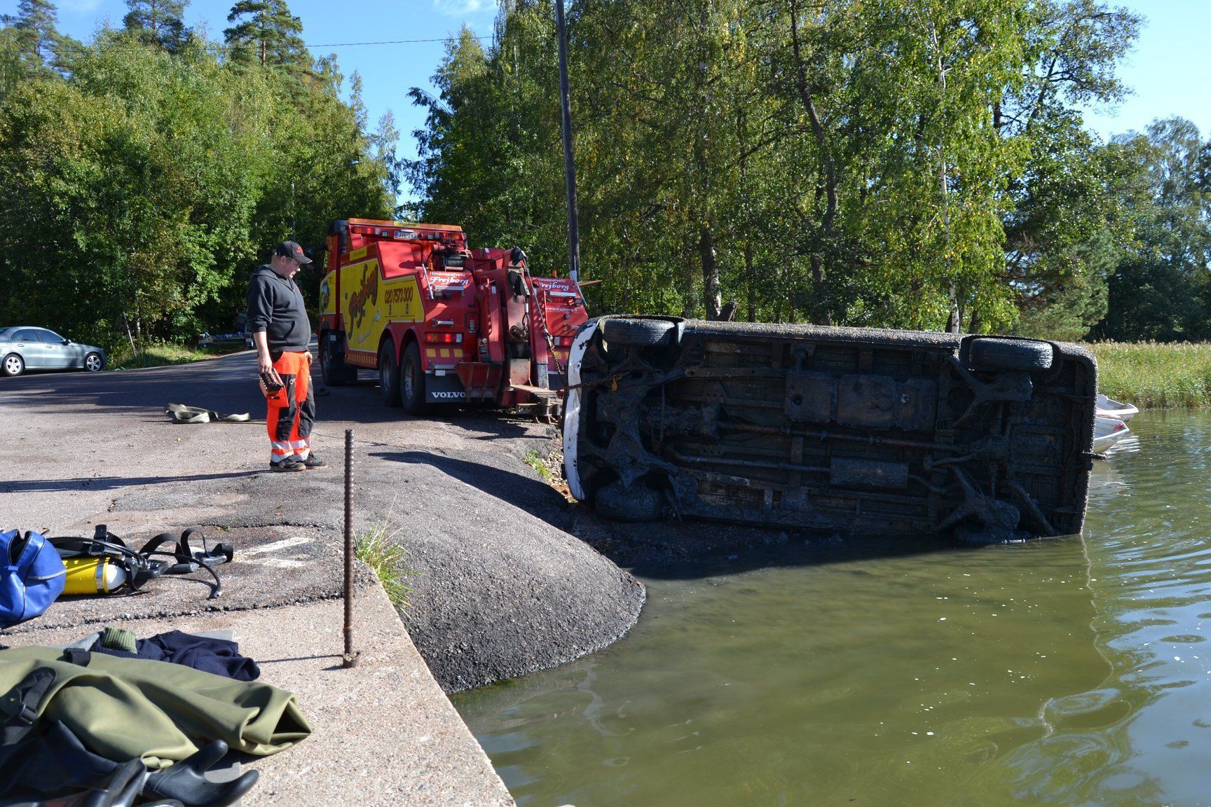Kaatunut musta ajoneuvo vedessä tien laidassa, pelastustyöntekijät ja hinausauto lähistöllä