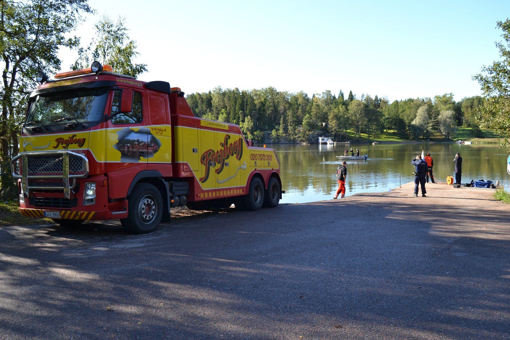 Punainen paloauto järvenrannan veneluiskalla, rannalla ihmisiä ja kartioita.