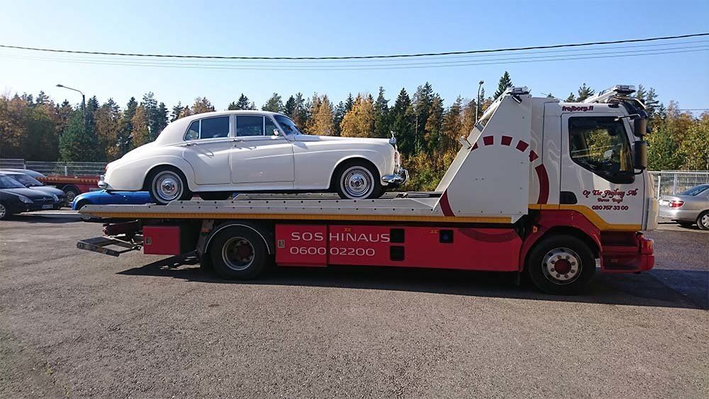Klassinen valkoinen auto punaisessa hinausautossa pysäköitynä ulkona puita taustalla