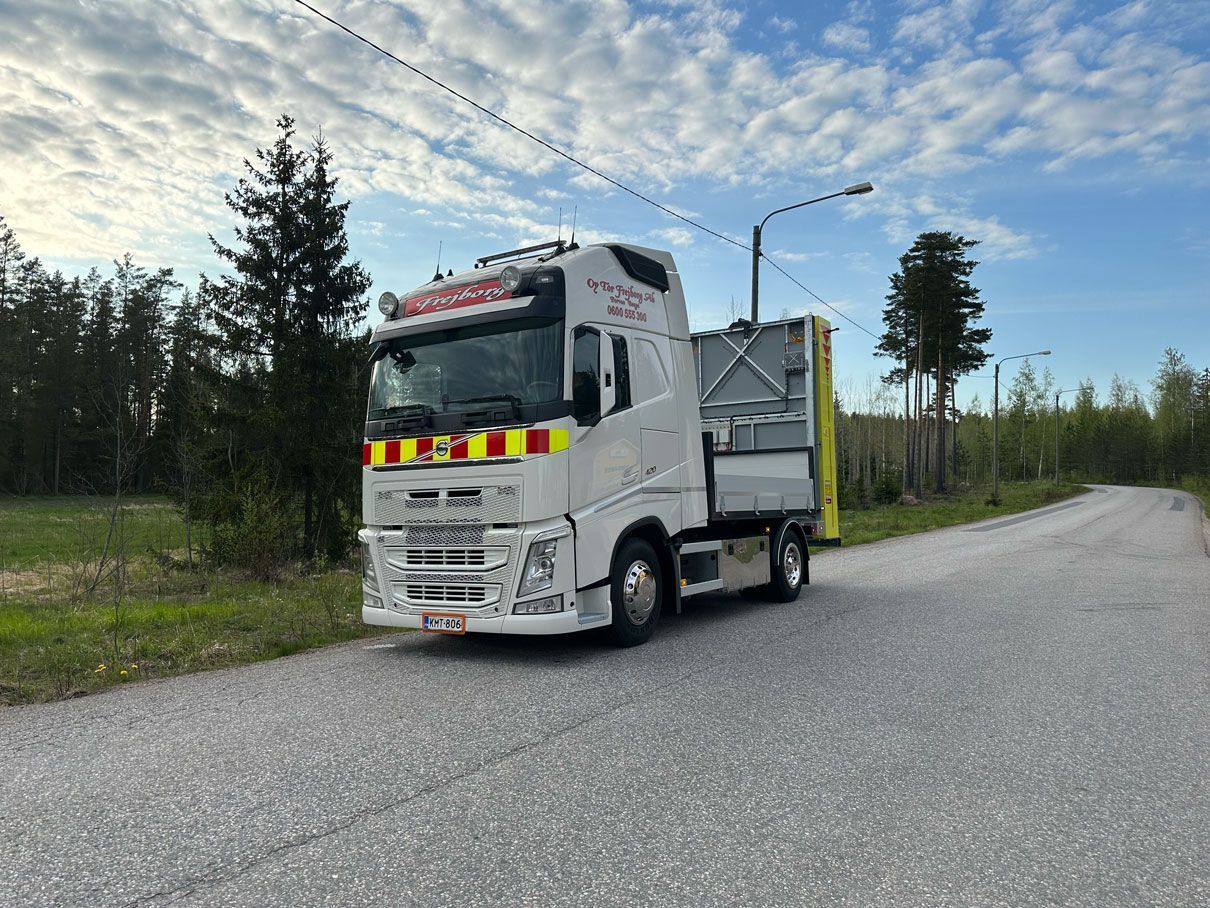 Valkoinen hinausauto pysäköitynä soratielle metsän viereen pilvisen taivaan alla