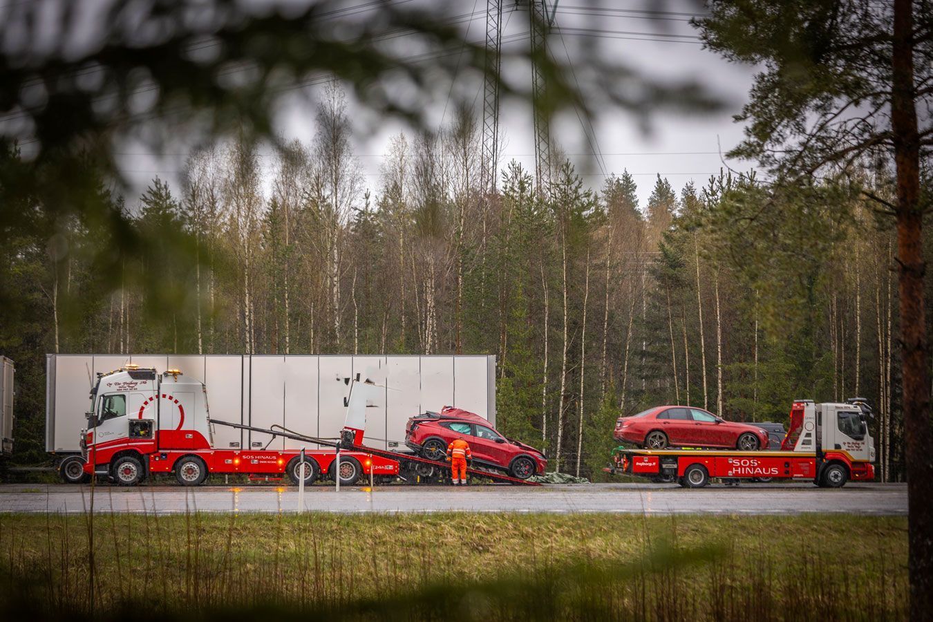 Punaiset hinausautot vetävät valkoisia perävaunuja metsäisen pellon reunalla olevaa tietä pitkin.