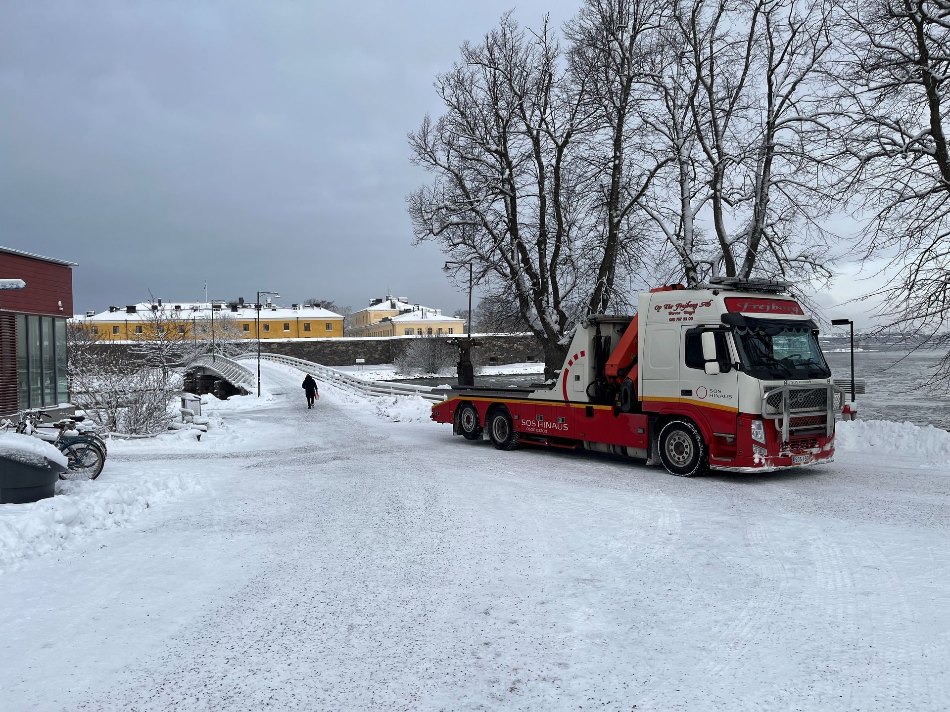 Punainen hinausauto lumisella tiellä paljaiden puiden ja rakennusten lähellä harmaan talvitaivaan alla
