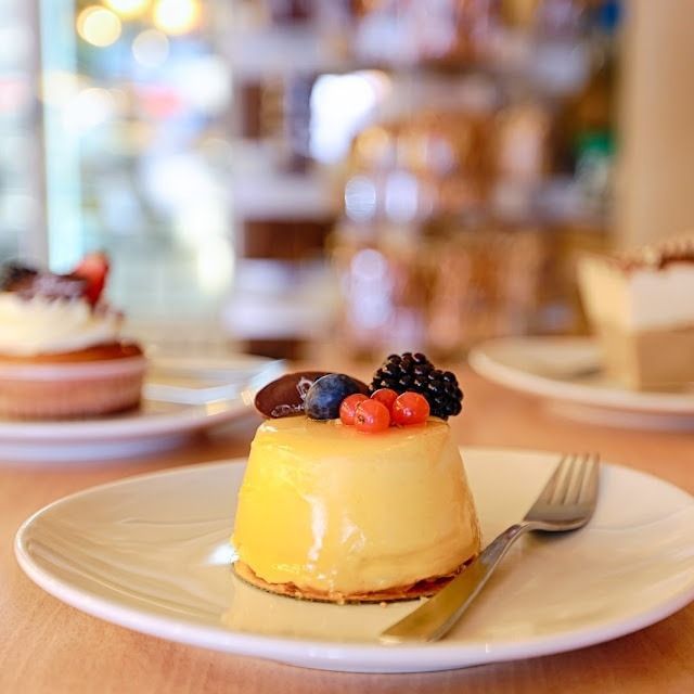 Un dessert jaune brillant garni de baies, présenté sur une assiette blanche avec une fourchette, d'autres pâtisseries se détachant floutement en arrière-plan.