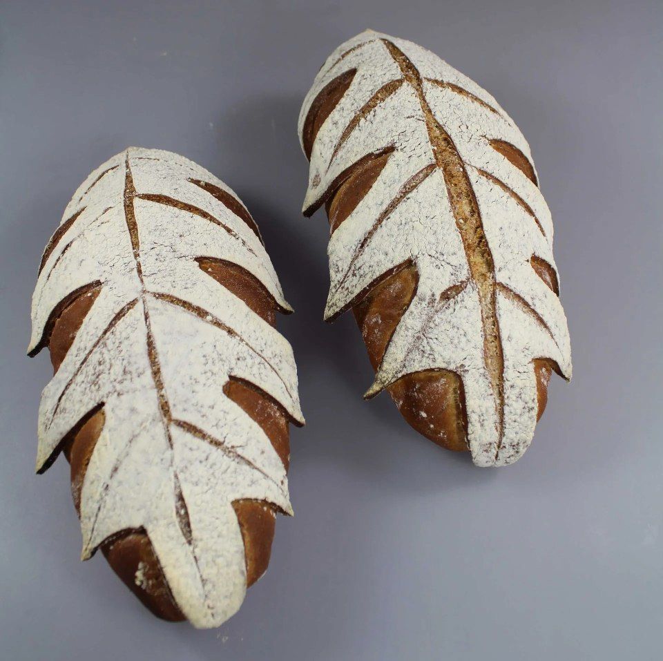 Deux miches de pain au levain artisanal, incisées en forme de feuilles et saupoudrées de farine, se détachent sur un fond gris uni.