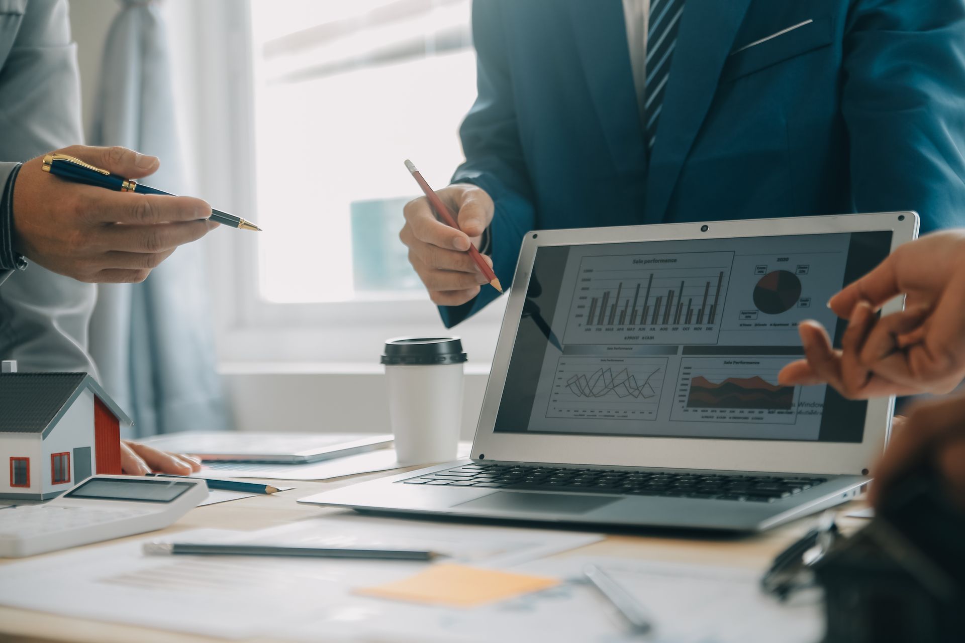Personas trajeadas revisan datos en una computadora portátil, discutiendo gráficos e información financiera. Cerca hay una maqueta de una casa.
