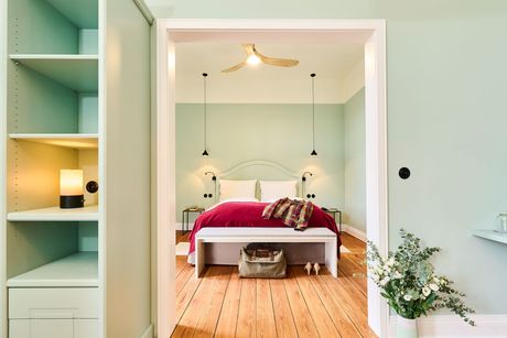 Helles Schlafzimmer mit rotem Bett, hellgrünen Wänden, Holzboden und eingebauten Regalen.